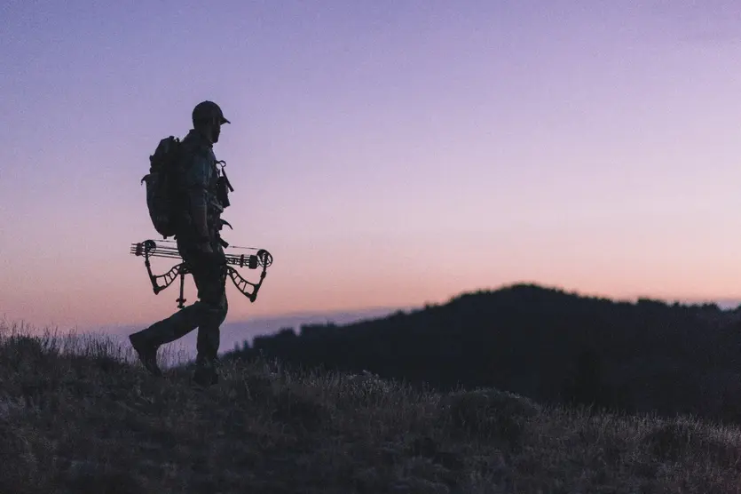 Archery hunter hiking with bow in hand on the mountain at dusk.